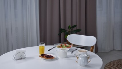 Shot of stylish living room with dining table, freshly prepared meals standing on the table, salad bowl, toasts with jam and glass of juice with coffee cup.