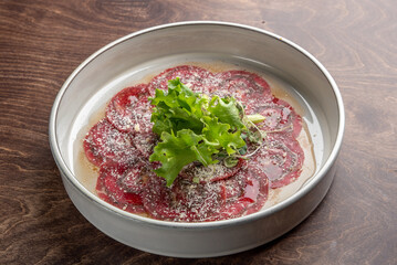 A beautifully arranged plate of beef carpaccio, featuring thinly sliced red meat garnished with fresh greens and a sprinkle of parmesan cheese, served in a stylish bowl on a wooden surface.