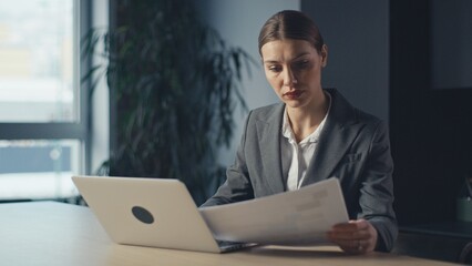 Businesswoman in a formal suit working at her desk, reviewing documents with a laptop in a modern office environment. Concept of professionalism, corporate responsibility, and focus