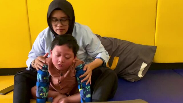 An Indonesian boy living with cerebral palsy is undergoing a therapy session, assisted by a female physiotherapist wearing a headscarf in a physiotherapy room surrounded by a soft mattress.