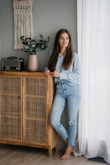 A young woman in a light blue shirt and jeans leans casually against a rustic cabinet, exuding a relaxed vibe in a cozy, minimalist room.