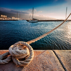 Close up view of a mooring rope secured to a harbor wall with boats floating in the background at sunset