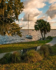 A sailboat rests on the water near a park bench and palm tree at Boathouse Riverfront Park, Palatka, Florida