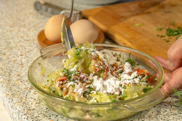 Chef mixes zucchini pulp grated on a grater, herbs, flour, paprika and chicken egg for making pancakes with a spoon in a glass plate. Close-up