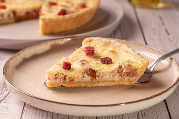 fatia de quiche lorraine em um prato de ceramica sob uma mesa de madeira clara e um quiche atrás...