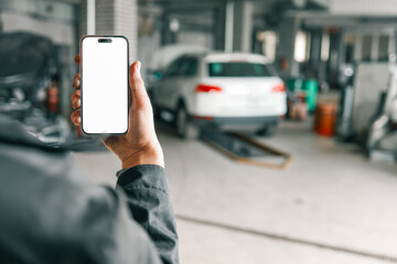 Hand holding a smartphone with a blank white screen in a car repair workshop, featuring blurred vehicles and garage equipment in the background