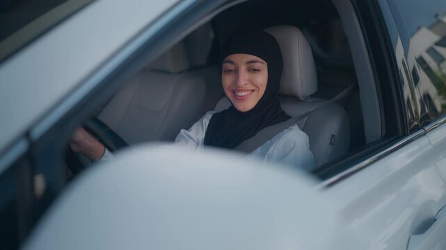 Middle Eastern young woman in hijab sitting in car. Smiling while looking at side mirrors and rotating steering wheel. Confident female carefully maneuvering white vehicle backwards in urban setting.