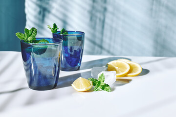 Summer refreshing lemonade drink with mint, ice and lemon slices on white surface and blue background. Fresh healthy cold lemon beverage. Infused water with lemon.