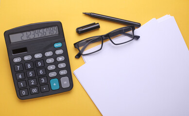 Black classic calculator with white paper sheets, eyeglasses, pen on yellow background. Business concept. Top view