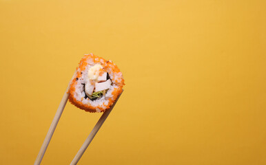 Chinese chopsticks holding sushi roll on yellow background