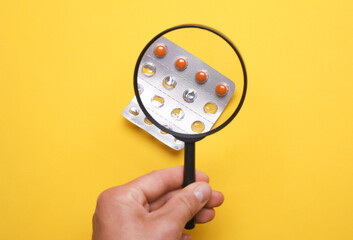 Hand with a magnifying glass examines blister pills on yellow background
