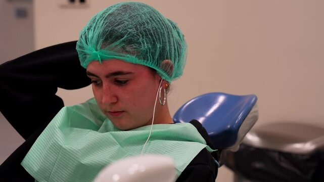 Young anxious patient sitting in dental chair listening to music waiting doctor wearing surgical cap