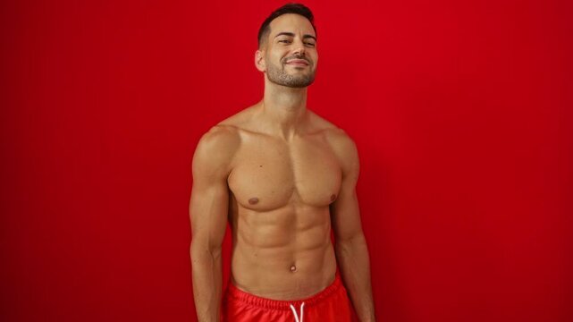 Swimsuit-clad young hispanic man shirtless and sexy blowing a kiss on air, expressing love and playfulness while looking lovely over isolated red background