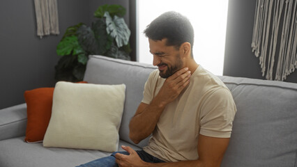 Fototapeta premium Young hispanic man with beard sitting on sofa at home rubbing neck in discomfort, surrounded by pillows and plants in a cozy living room interior.