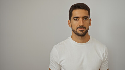 Handsome hispanic man with a beard stands over an isolated white background wearing a white shirt, exuding a sense of confidence and attractiveness in the simplicity of his appearance.