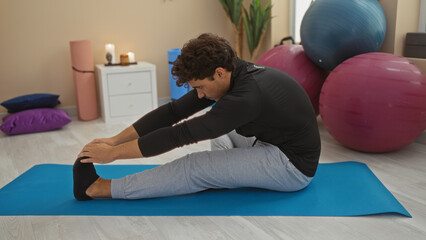 Young man stretching on a yoga mat in a gymnasium surrounded by exercise balls and fitness equipment, maintaining balance and form indoors.