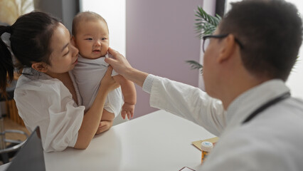 Mother holding baby boy with male pediatrician in hospital clinic room, emphasizing caring family love in healthcare setting.