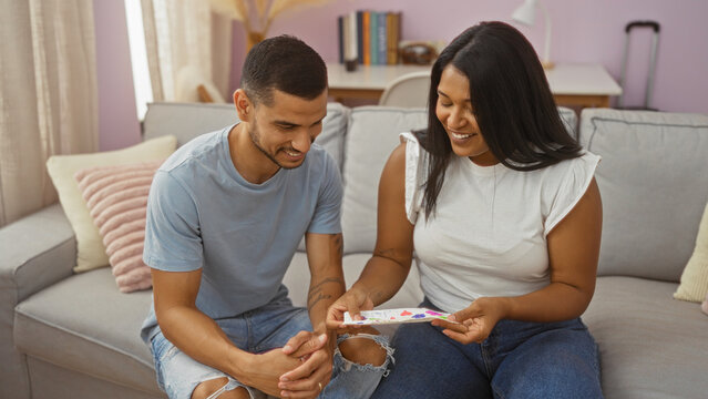 Couple sitting on a couch, smiling and sharing a moment while holding a colorful card in their living room showing affection and togetherness in a cozy indoor setting