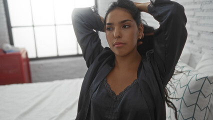 Young, brunette, woman in a bedroom setting, with a thoughtful expression, sitting on a bed,...