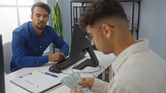 Two hispanic men working together in an office with one focused on paperwork and the other counting united states dollar bills, creating a professional business environment indoors