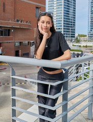 A young woman with dark hair and glasses on her head poses leaning on a railing, one hand on her face. She is wearing a black T-shirt and dark pants. In the background, modern urban style buildings