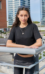 A young woman with long dark hair, standing by a railing, wears a black T-shirt and dark pants. She wears glasses on her head and a pendant. In the background, modern urban fashion buildings