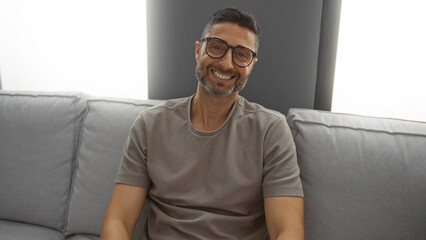 Young man with glasses smiling warmly indoors on a gray couch in a cozy living room setting...