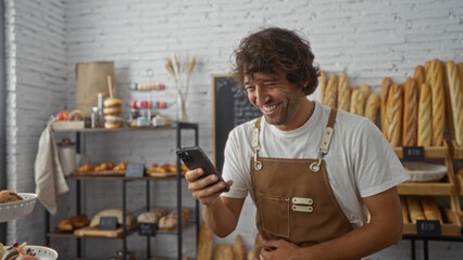 Young man in bakery wearing apron smiling at phone surrounded by bread and pastries in a cozy...