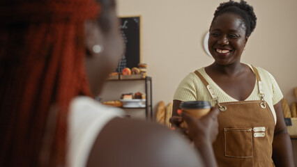 Two african american women interacting in a bakery with one as an employee in an apron and the other as a customer holding a coffee cup