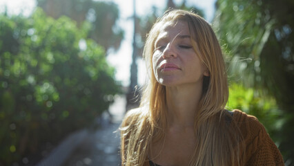 Woman enjoying sunlight in urban park, with eyes closed, blonde hair, beautiful smile, outdoors in city during sunny day, surrounded by greenery and peaceful ambiance