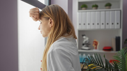 Woman leaning on a window looking tired in a hospital room, with shelves, binders, and medical equipment in the background, conveying a scene of workplace stress.