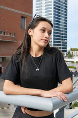 A young woman with long dark hair, leaning on a railing, wears a black T-shirt with a pendant of theatrical masks and sunglasses. In the background, a modern building and a red brick building. 