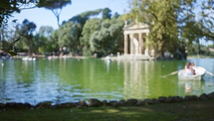 Summer tourists enjoy boating on serene lake in blurred villa borghese gardens, with lush green surroundings and classical architecture in rome, italy, under a clear blue sky.