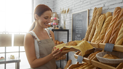 Young woman with red hair pleasantly smiling while inspecting freshly baked bread in a cozy bakery shop interior.