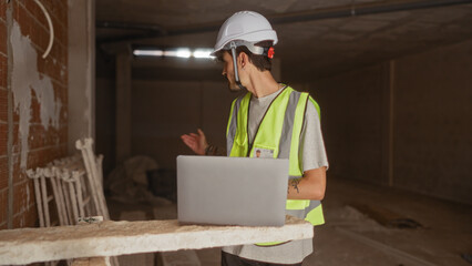 Young man working at a construction site with a laptop wearing a hardhat and reflective vest, in a partially built house, appearing focused on the work.