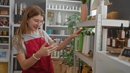 Young woman in glasses with blonde hair, wearing a red apron, uses her phone while shopping in a home decor store filled with beautifully arranged items on shelves.