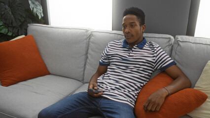 Young man sits relaxed on a couch holding a remote in a modern living room with stylish decor and cushions, capturing a serene indoor moment at home.