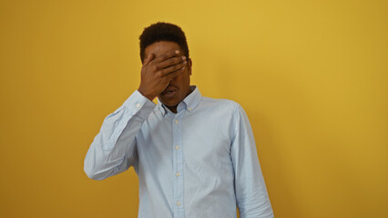 Young african american man in white shirt with hand on face standing against isolated yellow background looking contemplative and thoughtful.
