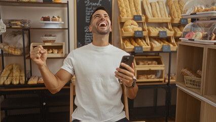 Young hispanic man smiling inside a bakery while holding a phone surrounded by fresh bread and pastries