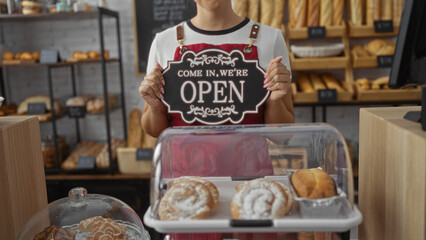 Woman holding open sign in bakery shop with pastries and bread in background