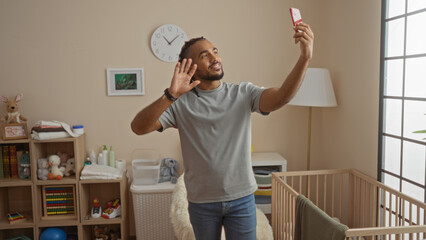 Young man with braids making video call in bedroom near cradle, waving and smiling warmly at smartphone, expressing connection in a comfortable home setting.