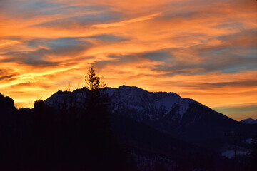 Golden hues of the sunset sky, blending vibrant shades of orange and yellow, cast a warm glow over the majestic High Tatras mountain peaks. Dark silhouettes of trees, dramatic contrast.