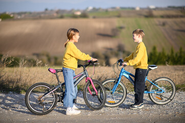 Fototapeta premium A boy and a girl race their bicycles through the countryside at sunset, laughing and chasing each other. The golden light and peaceful surroundings create a joyful, carefree moment of childhood advent