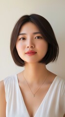 Fototapeta premium Portrait of a beautiful young asian woman with short dark hair, wearing a white sleeveless top and a delicate necklace, posing confidently against a minimalist white background
