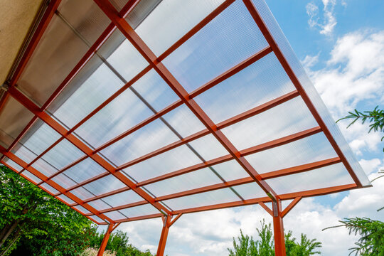 Transparent polycarbonate roof on a modern wooden pergola structure in a backyard, providing shade and protection with a clear sky background.