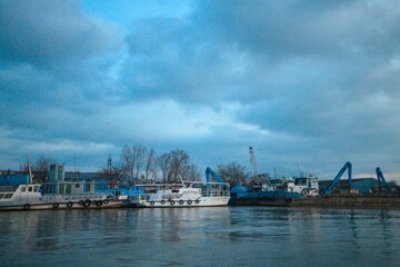 Fototapeta premium Boats rest quietly along the waterfront, surrounded by a vibrant mix of industrial buildings. The sky hangs low and dramatic, casting reflections on the water's surface as dusk approaches