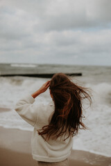 A young girl stands with her back to the camera against the backdrop of a turbulent sea, the wind blowing through her hair. She is wearing a white sweater.