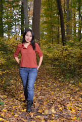 A beautiful young Asian woman in a reddish orange blouse and jeans walks through woods in fall