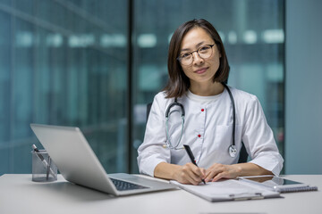 Portrait of an Asian woman inside a clinic office at her workplace. Female doctor behind paperwork...