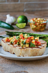 grilled swordfish topped with mango salsa served with asparagus on a white plate